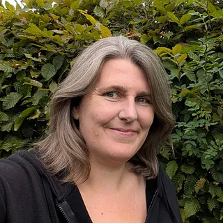 Portrait von Gesche Schaar. Sie steht vor einer Hecke und lächelt in die Kamera. Sie trägt die Haare schulterlang und offen. Sie trägt einen schwarzen Pulli.